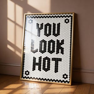 May include: Framed art print with the text "YOU LOOK HOT" in bold black letters. The background is a mosaic of white and black hexagonal tiles. The frame is gold-colored, and the artwork is leaning against a wall.