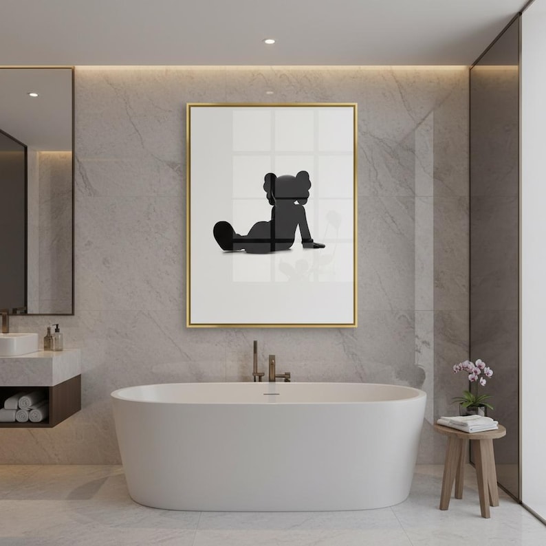May include: A framed art print featuring a black silhouette of a seated figure with a distinctive head shape. The artwork is set against a white background within a gold frame. The print is displayed in a modern bathroom setting with a white bathtub.