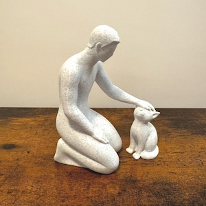 May include: A speckled white sculpture of a person kneeling and petting a small cat. The figures are detailed with a speckled pattern, creating a textured appearance. The sculpture is set on a wooden surface.