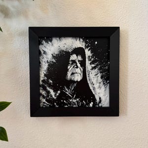May include: A framed black and white portrait of a stern-faced figure in a hooded cloak. The artwork features a dramatic, expressive style with bold brushstrokes and a focus on the subject's face. The black frame adds a clean, modern touch.