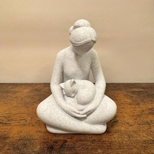May include: A speckled white sculpture of a seated figure cradling a curled-up cat. The figure has a bun hairstyle and is gazing downward. The cat is nestled in the figure's lap, creating a serene and comforting scene.