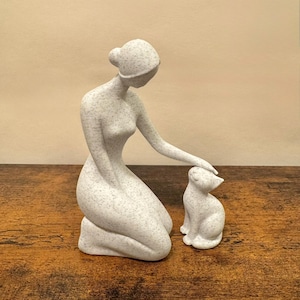 May include: A speckled white sculpture of a kneeling figure gently touching a seated cat. The sculpture has a smooth, minimalist design with a neutral color palette. The cat is small and has its head tilted up, looking at the figure.