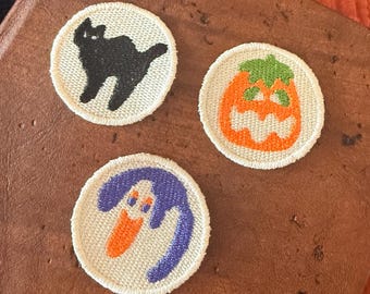 May include: Three circular embroidered patches with a cream-coloured background. One patch features a black cat, another displays an orange pumpkin with a green stem, and the third shows a purple ghost-like figure with an orange face.