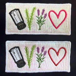May include: Two rectangular embroidered patches featuring a white background with a subtle iridescent sheen. Each patch displays a black and white salt shaker, a green sprig of rosemary, a purple lavender sprig, and a red heart outline.