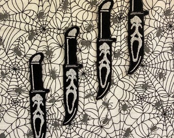 May include: Four embroidered patches shaped like knives with black handles and white blades featuring a stylised ghost face design. The patches are arranged in a diagonal line against a white fabric background printed with a black spider web pattern.