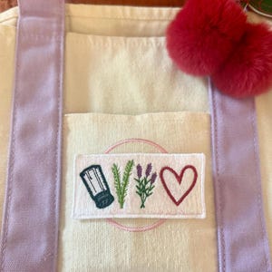 May include: A cream-coloured canvas tote bag with light purple straps and a rectangular patch featuring embroidered icons of a salt shaker, a sprig of rosemary, a sprig of lavender, and a red heart. A pair of fuzzy red pom-poms hangs from the strap.