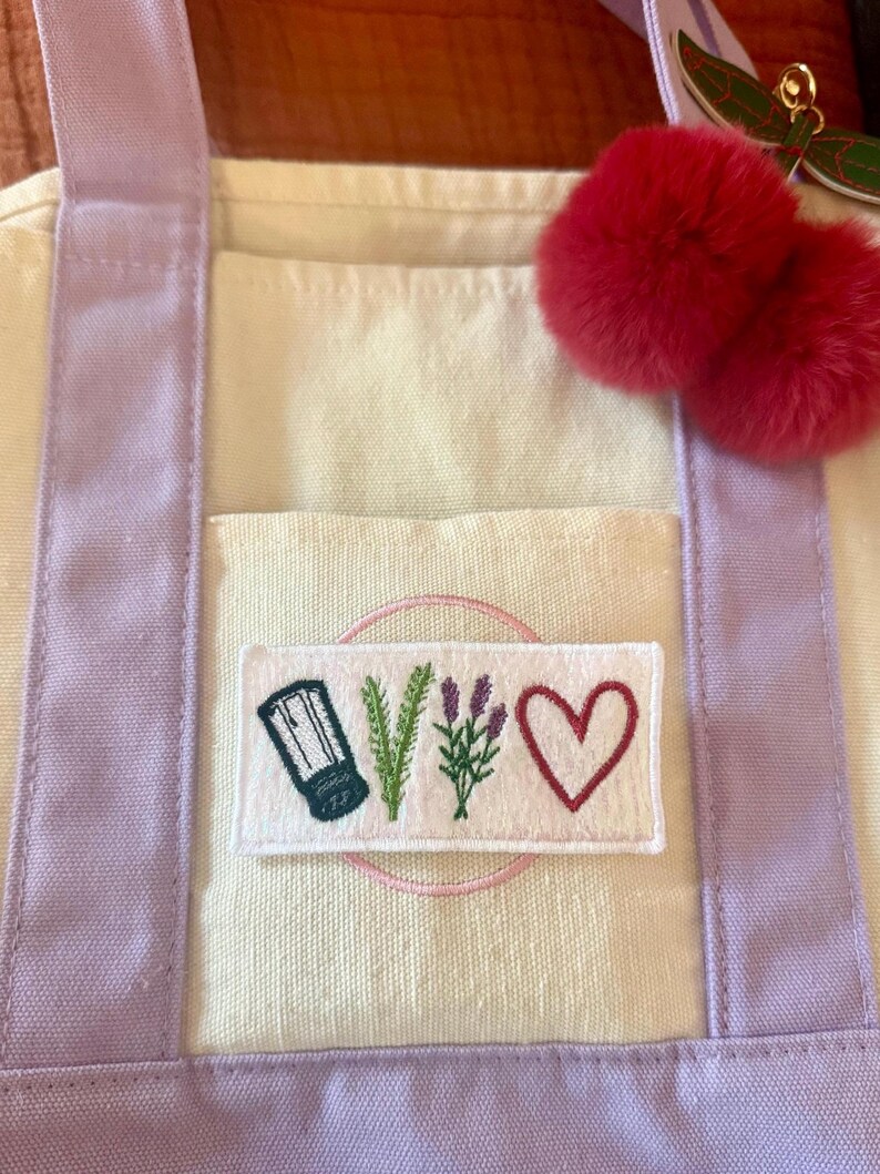 May include: A cream-coloured canvas tote bag with light purple straps and a rectangular patch featuring embroidered icons of a salt shaker, a sprig of rosemary, a sprig of lavender, and a red heart. A pair of fuzzy red pom-poms hangs from the strap.