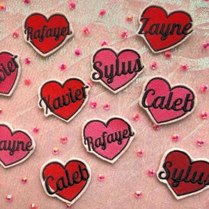 May include: A collection of heart-shaped embroidered patches in red and pink, each with a name in a cursive font. The patches are scattered on a pink surface with small, scattered pink beads. Names include Xavier, Caleb, and Zayne.