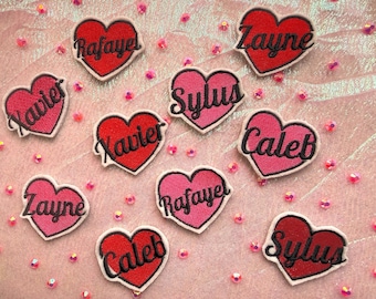 May include: A selection of heart-shaped embroidered patches in red and pink, each with a name in a cursive font. The patches are scattered on a pink surface with small, scattered pink beads. Names include Xavier, Caleb, and Zayne.
