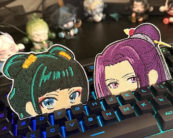 May include: Two embroidered patches depicting anime-style female characters with detailed facial features. One has green hair and blue eyes, the other has purple hair and a purple outfit. The patches are on a black keyboard.