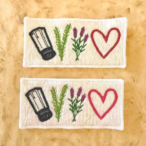May include: Two rectangular white embroidered patches featuring a black salt shaker, a green sprig of rosemary, a purple sprig of lavender, and a red heart outline. The patches have a white stitched border and a textured fabric base.