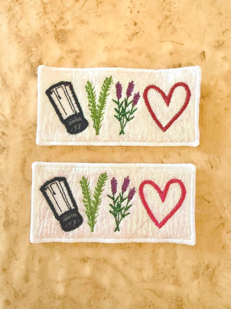 May include: Two rectangular white embroidered patches featuring a black salt shaker, a green sprig of rosemary, a purple sprig of lavender, and a red heart outline. The patches have a white stitched border and a textured fabric base.