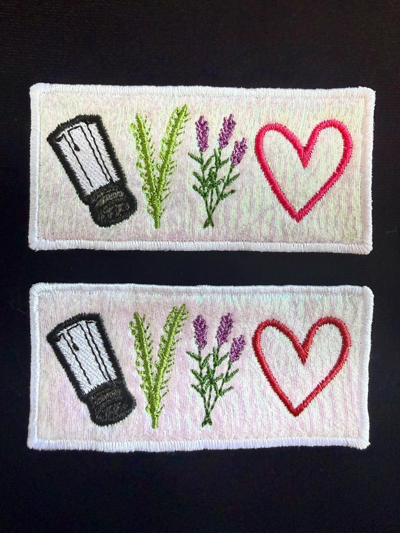 May include: Two rectangular embroidered patches featuring a white background with a subtle iridescent sheen. Each patch displays a black and white salt shaker, a green sprig of rosemary, a purple lavender sprig, and a red heart outline.