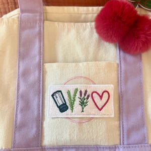 May include: A cream-coloured canvas tote bag with light purple straps and a rectangular patch featuring embroidered icons of a salt shaker, a sprig of rosemary, a sprig of lavender, and a pink heart. A pair of fuzzy red pom-poms hangs from the strap.