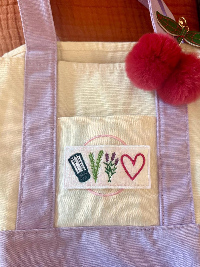 May include: A cream-coloured canvas tote bag with light purple straps and a rectangular patch featuring embroidered icons of a salt shaker, a sprig of rosemary, a sprig of lavender, and a pink heart. A pair of fuzzy red pom-poms hangs from the strap.
