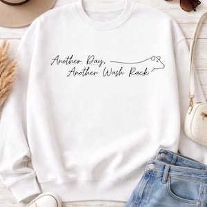 May include: White sweatshirt with the text "Another Day, Another Wash Rack" in a script font. The design includes a line drawing of a cow. The sweatshirt is paired with blue jeans, sunglasses, a purse, a hat, and sneakers.