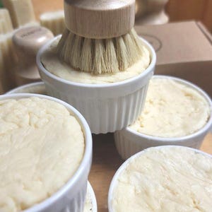 Könnte beinhalten: Nahaufnahme von mehreren weißen Auflaufförmchen, gefüllt mit einer cremigen, cremefarbenen Substanz. Eine Bürste mit Holzgriff und hellen Borsten ist über einem Auflaufförmchen positioniert. Weitere Seifen und eine Schachtel befinden sich im Hintergrund.