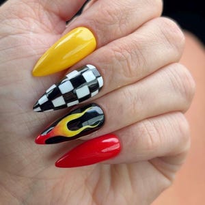 May include: Close-up of a hand with long, pointed fingernails. The nails are painted in various designs: solid yellow, a black and white checkered pattern, a flame design with red, yellow, and black, and solid red. The nails are glossy and well-manicured.