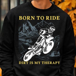 Peut inclure: Un sweat-shirt noir à col rond avec un motif blanc représentant une personne sur une moto tout-terrain dans un paysage montagneux. Le texte BORN TO RIDE est imprimé en jaune au-dessus et DIRT IS MY THERAPY est imprimé en jaune en dessous.