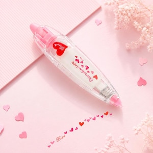 Puede incluir: Un dispensador de cinta correctora de pl&aacute;stico rosa y transparente con dise&ntilde;o de coraz&oacute;n. El dispensador tiene la inscripci&oacute;n "Decoration Tape" y dispensa una l&iacute;nea de peque&ntilde;os corazones rojos. La palabra "Love" tambi&eacute;n est&aacute; impresa en la cinta.