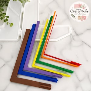 May include: A set of six L-shaped plastic alignment tools in brown, blue, purple, yellow, green, and orange, arranged in a nested stack. Each tool features a small rectangular tab at the end of one arm, positioned in front of a white plastic case.