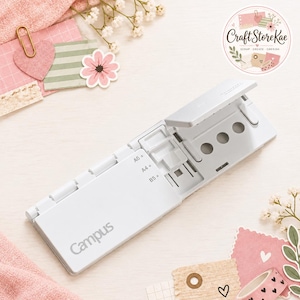 May include: A white Campus brand paper punch tool featuring a hinged design with adjustable settings for A6, A4, and B5 paper sizes. The device includes a series of circular punch holes and a clear plastic guide for alignment on the top surface.