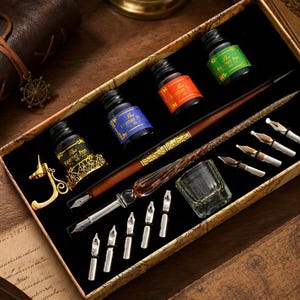 K&ouml;nnte beinhalten: Ein Kalligraphie-Set in einer rechteckigen Schachtel mit einem braunen Holzstift mit Metallfeder, einer goldfarbenen Stiftablage, einem klaren Glastintenfa&szlig; und vier kleinen Tintenflaschen mit Etiketten, die The Feather Pen in Schwarz, Blau, Rot und Gr&uuml;n tragen.