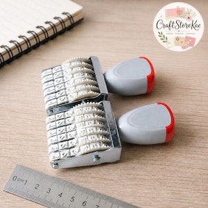 May include: Two side-by-side rolling date stamps with metallic frames, white plastic character wheels, and grey handles with red accents. A metal ruler is positioned below the stamps, showing a measurement scale in centimeters.