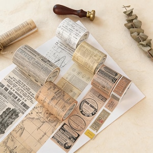 Puede incluir: Varios rollos de cinta adhesiva washi decorativa con diseños de periódicos y mapas antiguos. La cinta es en tonos marrones, beige y blancos. También hay un sello de madera y un papel enrollado.