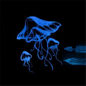 Puede incluir: Una ilustraci&oacute;n azul brillante de tres medusas sobre un fondo negro, con una medusa central grande y dos m&aacute;s peque&ntilde;as cerca. A la derecha, se ven dos plumines de pluma estilogr&aacute;fica distintos, tambi&eacute;n representados en un azul luminoso.