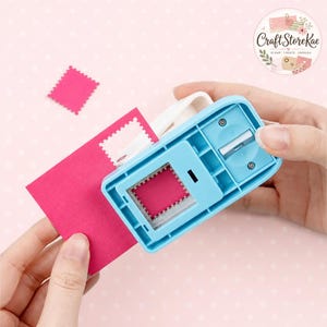 May include: A light blue paper punch tool with a rectangular opening, used to create a square shape with scalloped edges from a sheet of bright pink cardstock. The tool features a white lever mechanism and a metal cutting blade inside.
