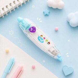 Puede incluir: Un dispensador de cinta correctora transparente y azul con un dise&ntilde;o de personaje morado. El dispensador tiene la palabra "pets" impresa, junto con varios personajes de dibujos animados. El fondo es azul claro con elementos decorativos.