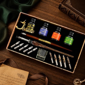 K&ouml;nnte beinhalten: Ein Kalligraphie-Set in einer rechteckigen Schachtel mit einem braun-goldenen F&uuml;llfederhalter, vier kleinen Tintenf&auml;ssern mit Etiketten der Aufschrift The Feather Pen, einer goldenen Stiftablage, einem kleinen Glas-Tintenfass und zehn silbernen Federn.