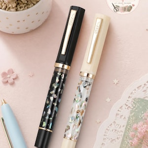 May include: Two fountain pens with gold-toned clips and caps, one in black and one in cream. The barrels are transparent and filled with iridescent, multi-colored shell fragments. Each cap is engraved with the text JINHAO in a vertical orientation.