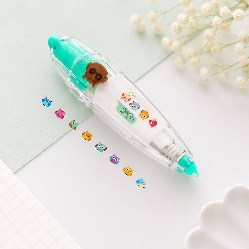 Puede incluir: Un dispensador de cinta correctora de pl&aacute;stico transparente con una tapa verde azulado y adornos en forma de b&uacute;ho. El dispensador tiene un cuerpo blanco con la palabra "Owls" impresa y est&aacute; decorado con pegatinas de b&uacute;hos coloridos.