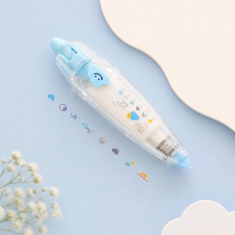 Puede incluir: Un dispensador de cinta correctora transparente, azul y blanco, con un dise&ntilde;o de nube. El dispensador tiene una tapa azul y un rollo de cinta blanco. Peque&ntilde;as pegatinas con tem&aacute;tica meteorol&oacute;gica est&aacute;n dispersas alrededor del dispensador sobre una superficie azul claro.