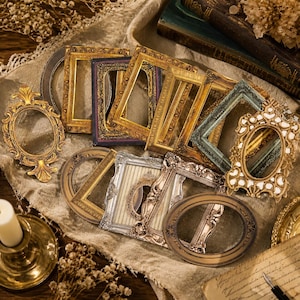 Può includere: Una collezione di cornici decorative in stile vintage. Le cornici sono principalmente dorate, con alcuni accenti argentati e scuri. Sono disposte su un tessuto strutturato di colore neutro, suggerendo un tema artigianale o decorativo.