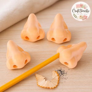 May include: Three peach-colored nose-shaped pencil toppers made of smooth material. One topper is attached to a yellow pencil, which has a small pile of wood shavings nearby. The design features realistic nostrils and a bridge, creating a distinct shape.