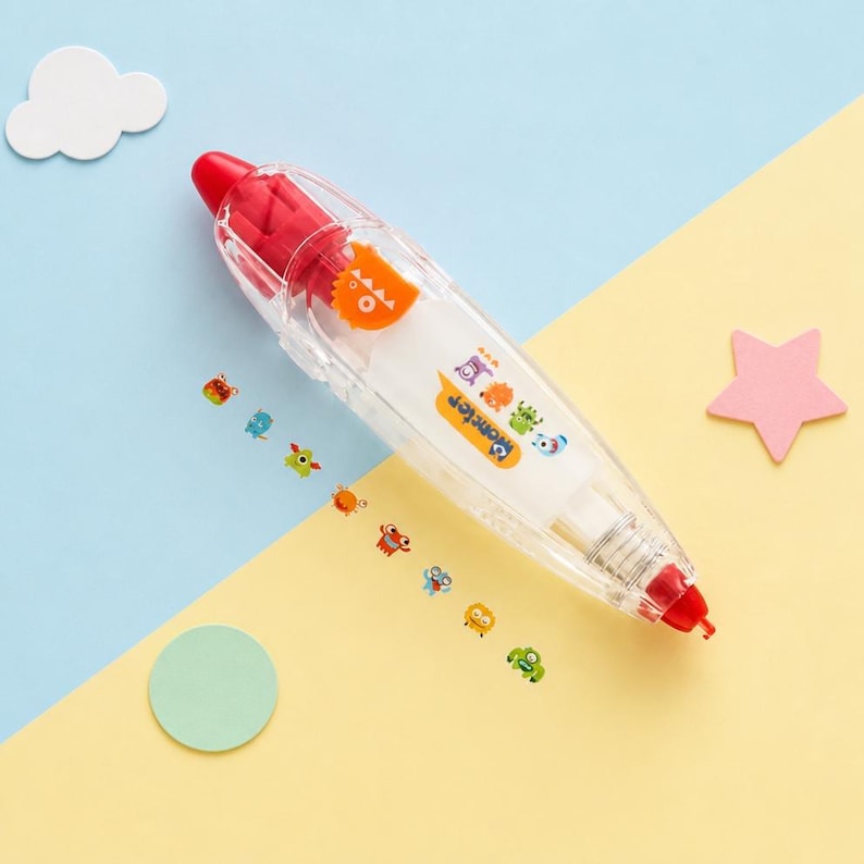 Puede incluir: Un dispensador de cinta correctora transparente con detalles rojos y un dise&ntilde;o de monstruo de dibujos animados. El dispensador tiene una punta roja y un monstruo naranja en el lateral. Peque&ntilde;as pegatinas de monstruos est&aacute;n dispersas.