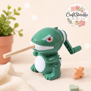 May include: A green dinosaur-shaped manual pencil sharpener with a white belly and a crank handle on the side. A wooden pencil is inserted into the dinosaur's mouth. The sharpener features a logo that reads Craft Store Kae, Scrap, Create, Cherish.