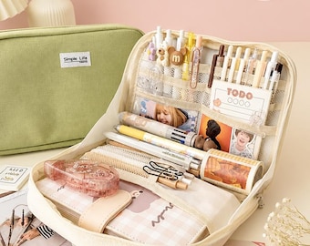 May include: A sage green rectangular pencil case with a Simple Life label, shown open to reveal a cream-colored interior filled with various pens, a TODO notepad, a roll of patterned tape, a pink correction tape, and a plaid-covered notebook.