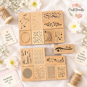 May include: A collection of rectangular wooden rubber stamps featuring various black ink designs including constellations, stars, shooting stars, floral wreaths, musical notes, geometric patterns, and moon phases, arranged on a light fabric surface.