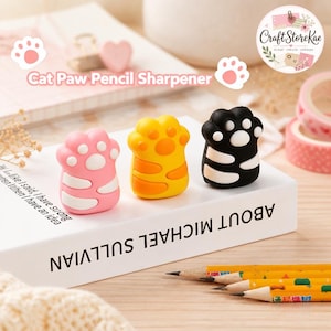 Puede incluir: Tres sacapuntas con forma de pata de gato en colores rosa, amarillo y negro. Cada sacapuntas presenta un diseño de almohadilla redondeada con detalles en blanco o naranja. Están alineados sobre un libro blanco con el texto ABOUT MICHAEL SULLVIAN.