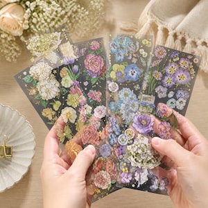 Könnte beinhalten: Eine Person hält mehrere transparente Blumenaufkleber. Die Aufkleber zeigen eine Vielzahl farbenfroher Blumen, darunter Rosen, Stiefmütterchen und Hortensien, mit goldenen Akzenten auf dunklem Hintergrund. Die Aufkleber sind fächerförmig angeordnet.