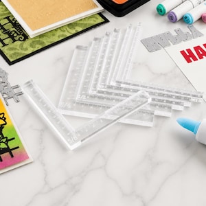 Könnte beinhalten: Ein Set aus transparenten, L-förmigen Acryl-Linealen mit aufgedruckten Bruchteil-Maßen in Zoll. Diese durchsichtigen Hilfsmittel dienen der präzisen Ausrichtung und Abstandsbestimmung bei Papierbastelprojekten in verschiedenen Größen.