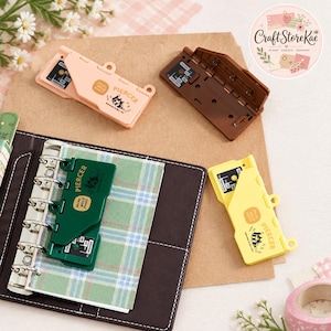 May include: A collection of four plastic mini hole punchers in light pink, dark brown, dark green, and yellow. Each puncher features the text PIERCER and a small graphic. One green puncher is attached to a six-ring binder with plaid paper.
