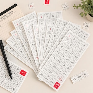 May include: A collection of white sticker sheets featuring black and red printed calendar dates and days of the week. The stickers are arranged in vertical strips and individual squares, with a black pen resting on one sheet and loose date labels scattered.