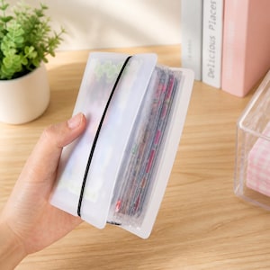 May include: A clear plastic card holder with a black elastic band closure. The holder is filled with various cards, visible through the translucent material. The holder is being held in a hand, with a wooden surface in the background.