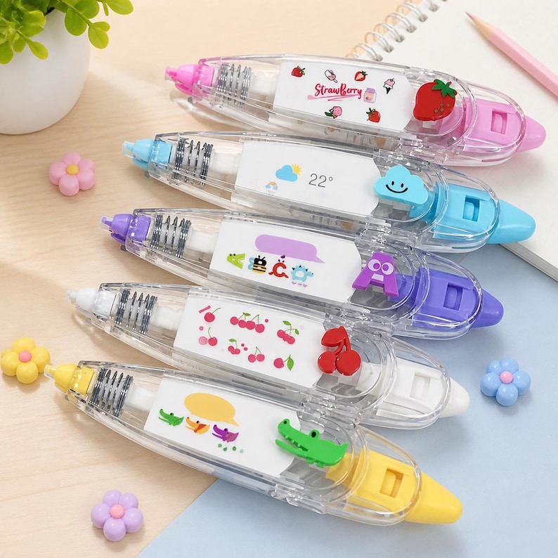 Puede incluir: Una colecci&oacute;n de dispensadores de cinta correctora transparentes con acentos coloridos. Cada dispensador presenta un dise&ntilde;o diferente, incluyendo fresas, un tema meteorol&oacute;gico, insectos, cerezas y un cocodrilo. Los dispensadores est&aacute;n dispuestos sobre una superficie azul claro.