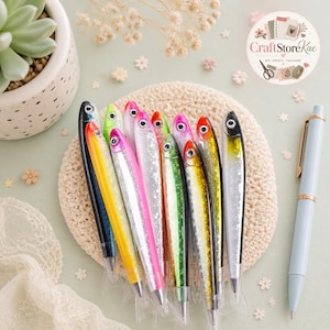 May include: A collection of ballpoint pens shaped like fish, featuring various colours including black, yellow, pink, green, and silver with glittery scales. Each pen has a realistic fish eye and a tail-shaped cap, arranged together in a cluster.